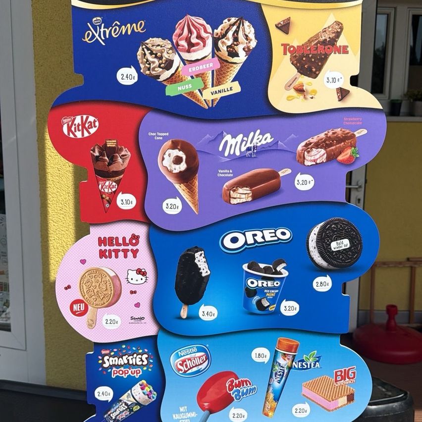 Ein Menübrett zeigt verschiedene Eiswaffeln und Eis am Stiel mit Preisen, darunter Kit Kat, Toblerone und Hello Kitty Eis am Stiel. Andere Artikel sind Oreo Eis am Stiel, Smarties, Nestle Schoeller und Bum Bum.