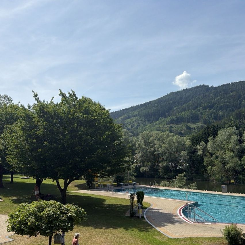 Ein Freibad mit blauem Himmel und einem Bergrücken im Hintergrund. Der Pool ist von Bäumen und Gras umgeben.