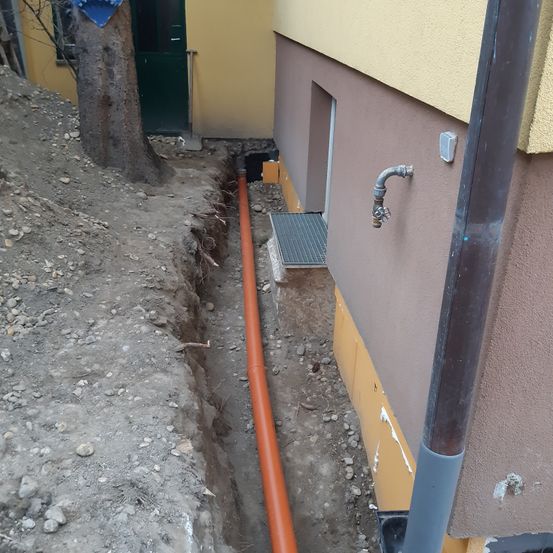 Ein neben einem Gebäude gegrabener Graben mit einer orangefarbenen und einer grauen Rohrleitung an der Wand. Eine Schaufel ist in der Nähe.