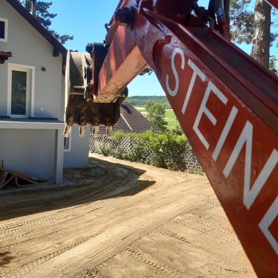 Ein roter Bagger mit der Aufschrift STEINE steht auf einem Erdweg mit einem Haus im Hintergrund.