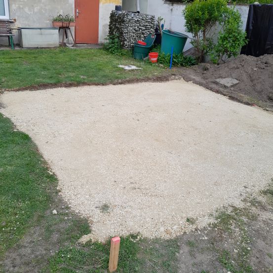 Ein Hinterhof mit einem frisch verlegten Kiesweg, umgeben von grünem Gras und Sträuchern. Ein Holzpfosten und einige Eimer sind ebenfalls vorhanden.