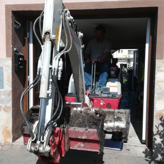 Ein Mann bedient einen roten und weißen Bagger, teilweise in einer Garage mit einer Steinmauer. Der Bagger ist mit Erde gefüllt.