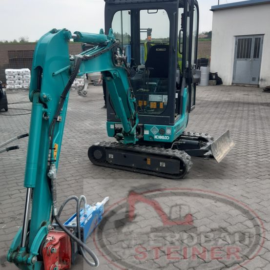Ein türkisfarbener Kobelco-Bagger steht in einem Außenbereich, mit einem Gebäude und Reifen im Hintergrund.