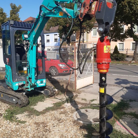 Ein türkisfarbener und schwarzer Bagger verwendet einen roten Bohrer, um ein Loch in den Boden zu bohren, mit einem eingezäunten Bereich und einem roten Auto dahinter.