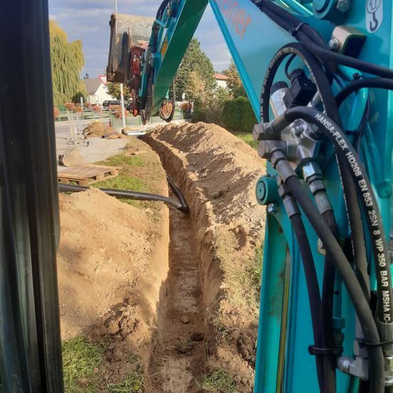 Ein blauer Bagger gräbt eine Rinne in einem erdenen Bereich mit Gras und einer Straße in der Nähe. Der Bagger ist mit verschiedenen Schläuchen und Kabeln verbunden.
