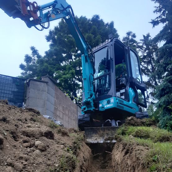 Ein blauer Bagger gräbt ein Loch auf einem Erdhügel mit einer Betonmauer im Hintergrund.