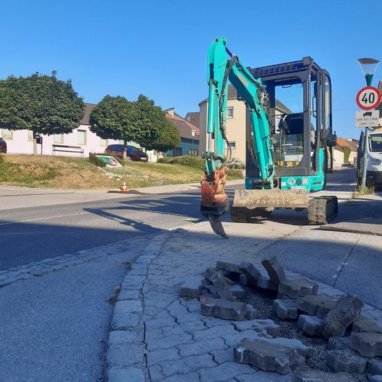 Ein grüner Bagger steht an einer Straßenecke und entfernt Betonpflaster. In der Nähe befinden sich Bäume, eine Bank und ein geparktes Auto. Ein Geschwindigkeitsschild ist sichtbar.
