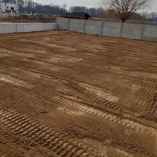 Ein Baustelle mit frisch umgegrabenem Boden, Reifenspuren, umgeben von Betonmauern und einem Haus im Hintergrund.