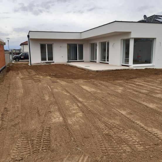 Ein modernes weißes Haus im Bau mit großen Glastüren und Fenstern ist von einem Erdhof umgeben. Ein geparktes Auto und ein Zaun sind in der Nähe.