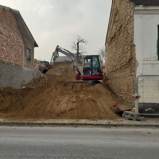 Ein roter und weißer Bagger gräbt Erde auf einer Baustelle aus, neben einem Ziegelgebäude und einem großen Erdhaufen.