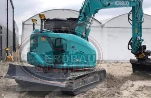 Ein grüner Bagger steht im Sand, dahinter ein Gebäude und eine Glasstruktur.