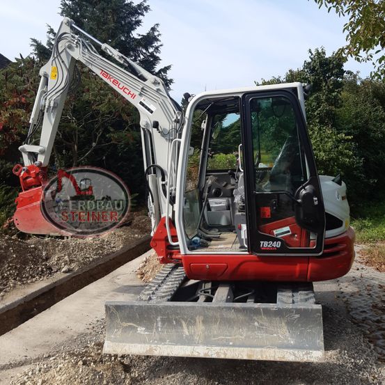 Ein Takeuchi TB240 Bagger steht auf einer Kiesfläche, im Hintergrund befinden sich Bäume und Sträucher. Der Bagger ist rot und weiß mit offener Tür.