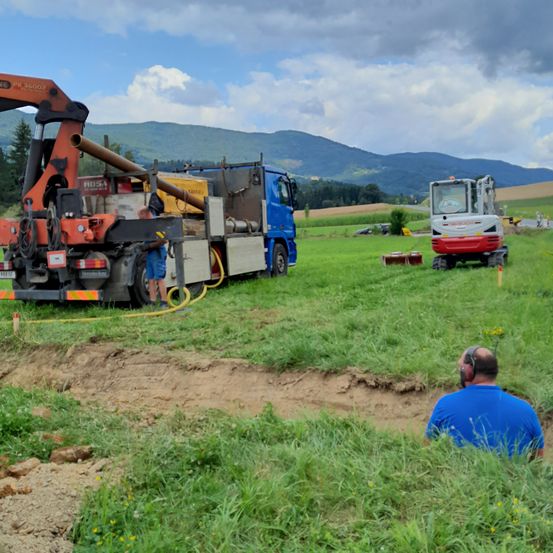 Eine Baustelle in einem grasigen Feld mit Bergen im Hintergrund. Zwei große Baumaschinen sind geparkt. Ein Mann in einem blauen Shirt sitzt und trägt Kopfhörer.