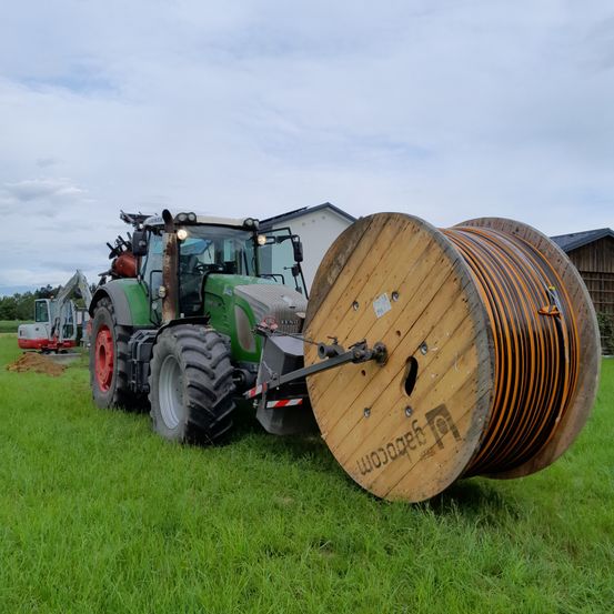 Ein grüner Traktor mit einer großen Kabelspule am Heck steht auf einem Grasfeld. Dahinter sind ein Gebäude und Maschinen zu sehen.