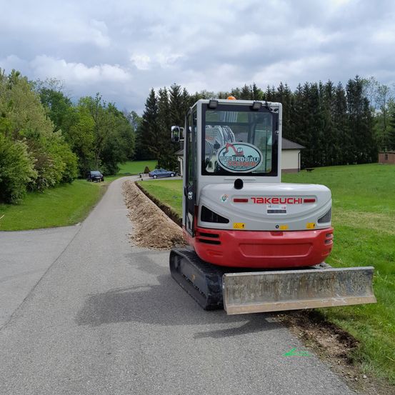 Ein roter und weißer Takeuchi-Bagger steht am Straßenrand mit einer grünen Grasfläche. Bäume und ein Haus sind im Hintergrund zu sehen.