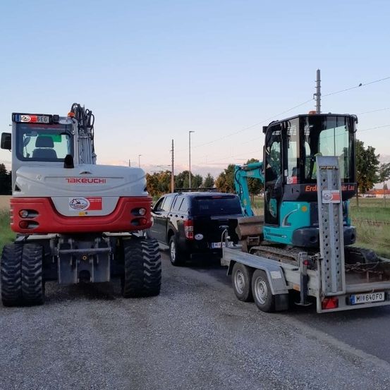 Ein Takeuchi-Traktor steht neben einem schwarzen SUV auf der Straße. Ein Anhänger mit einer blau-schwarzen Maschine ist am SUV befestigt. Bäume und Stromleitungen sind im Hintergrund.