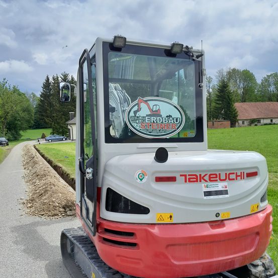 Ein roter und weißer Takeuchi-Bagger steht am Straßenrand, neben einer Erdstelle. Der Bagger hat ein Logo auf seiner Windschutzscheibe. Die Straße hat einen Grasrand mit Bäumen auf beiden Seiten.