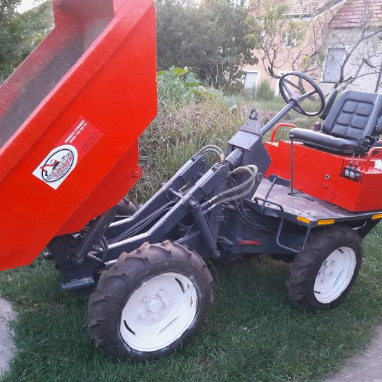 Ein roter Traktor mit Kippmulde steht auf einem Rasen. Er hat einen schwarzen Sitz und ein Lenkrad.