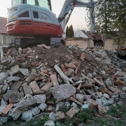 Ein roter und grauer Bagger steht auf einem großen Schutthaufen, wahrscheinlich für Abrissarbeiten. Dahinter befindet sich ein Gebäude mit einem Ziegeldach.
