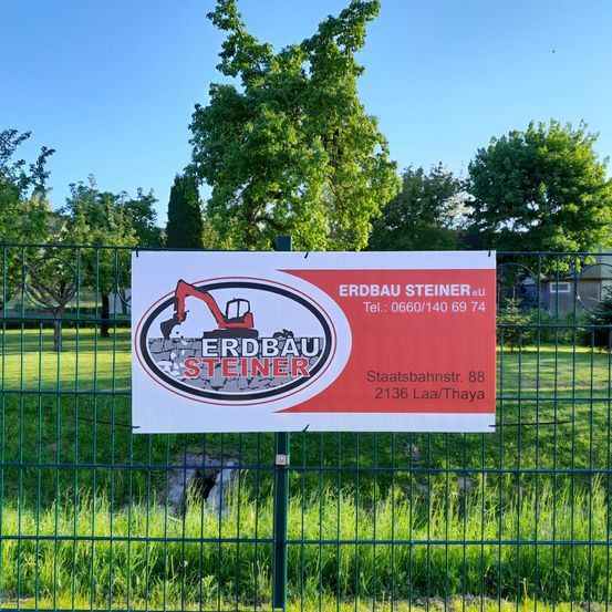 Ein rot-weißes Schild mit dem Text Erdbau Steiner ist an einem grünen Zaun befestigt. Es hat eine Telefonnummer und Adresse.