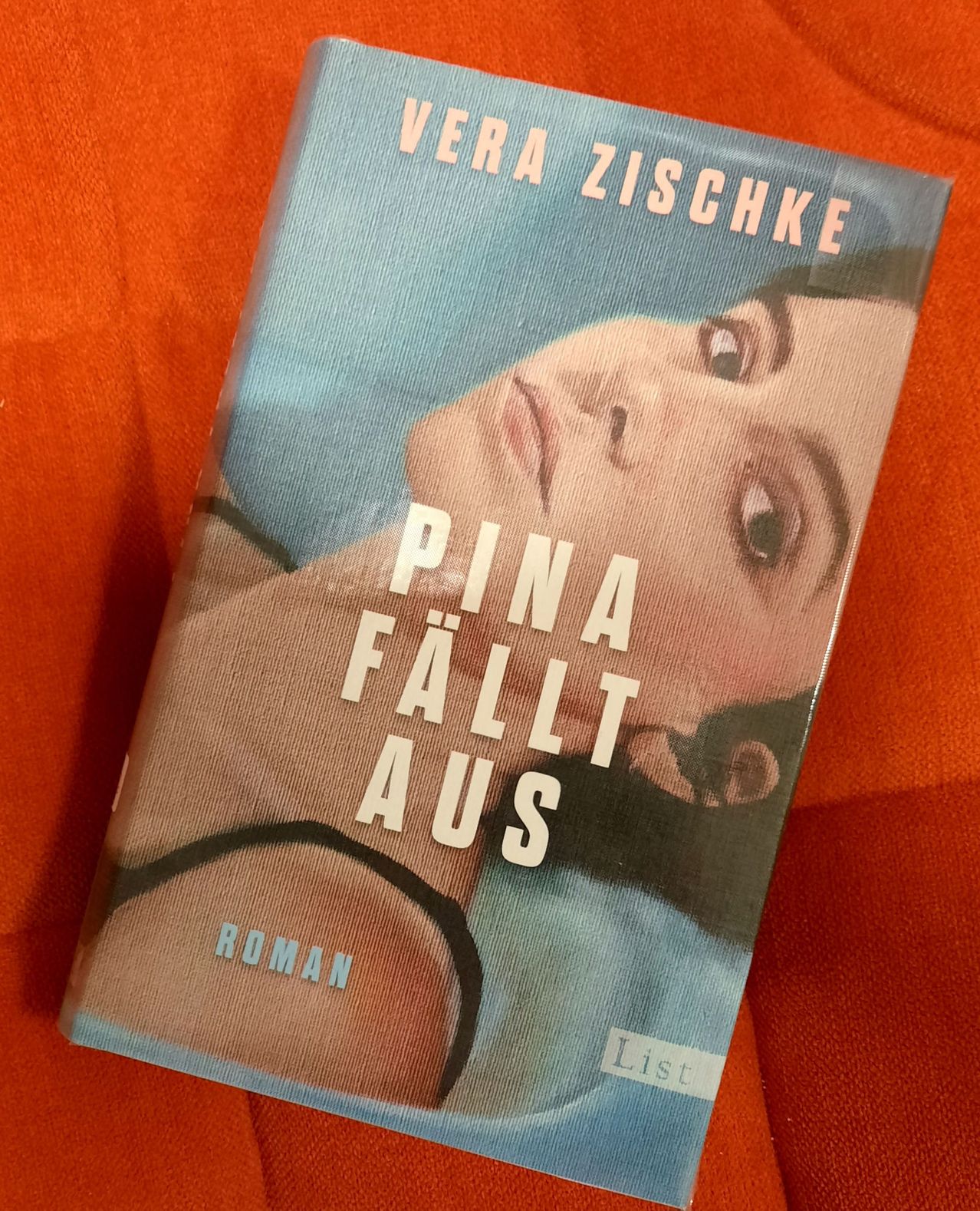 Ein Buch mit dem Titel 'Pina fällt aus' von Vera Zischke, das eine Frau auf dem Cover zeigt, die auf einem orangefarbenen Tuch liegt.