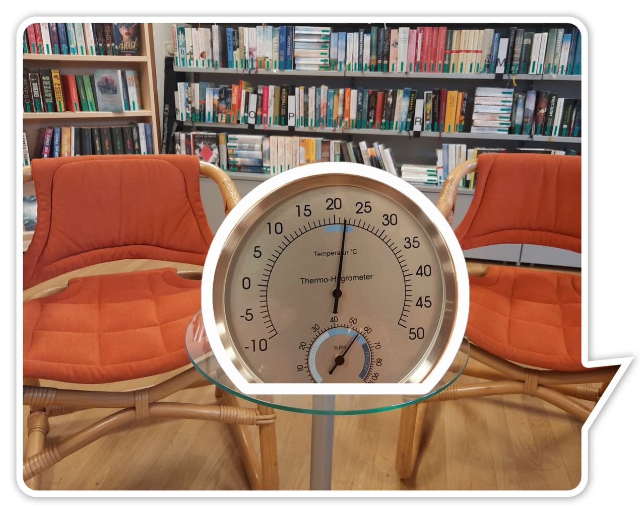 Ein runder Thermometer mit silbernem Rahmen steht auf einem Glastisch, umgeben von zwei orange gepolsterten Stühlen. Im Hintergrund befindet sich ein Bücherregal voller Bücher.