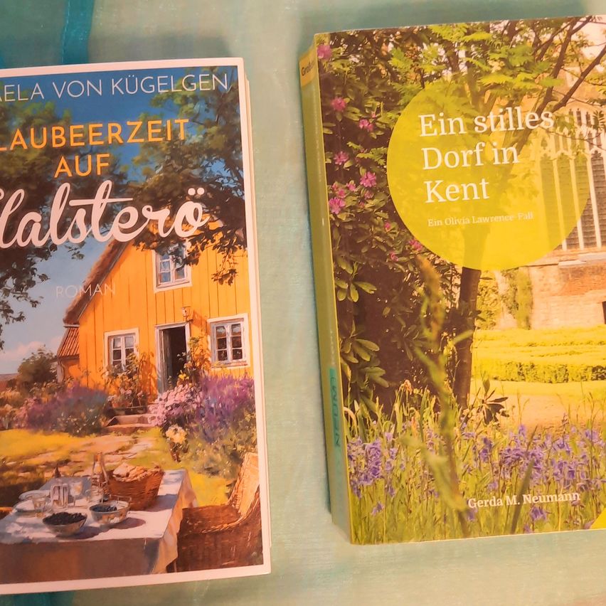 Two books on a table, one titled 'Laubeezeit auf Halstero' and the other 'Ein stilles Dorf in Kent'. The first book features a yellow house with a garden, while the second book shows a serene village scene.