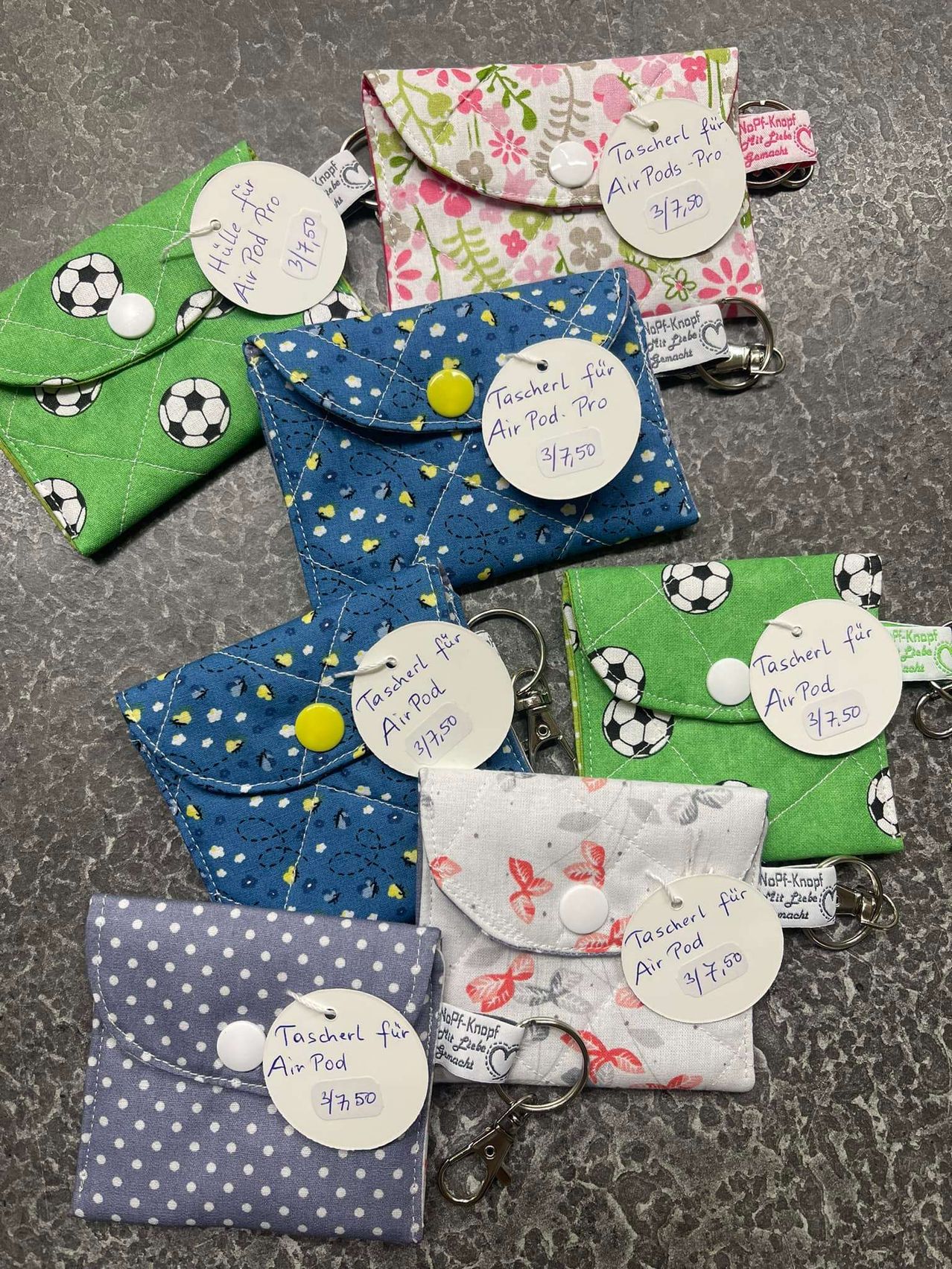 Mehrere kleine Taschen mit Schlüsselanhänger, jede mit einem Schild, das 'Tascherl fur Air Pod' und einen Preis von 3,75 Euro anzeigt. Sie sind in verschiedenen Farben und Designs, einige mit Fußballen.