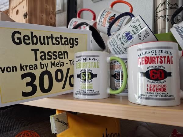 Ein Holzregal zeigt Tassen und Becher, darunter einige mit 'Geburtstags Tassen' Text. Ein gelbes Schild über den Tassen bietet einen 30% Rabatt. Die Tassen haben verschiedene Designs und Texte.
