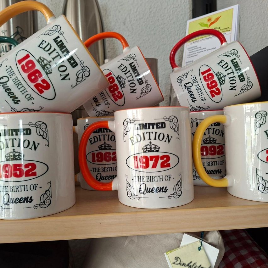 Ein Holzregal hält mehrere weiße Kaffeebecher mit roten und gelben Henkeln. Die Tassen haben die Aufschrift 'Limited Edition' und die Jahre 1952, 1962 und 1972.
