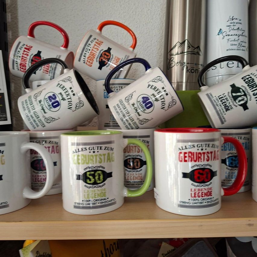 Ein Holzregal zeigt eine Vielzahl von Tassen mit verschiedenfarbigen Henkeln. Jede Tasse hat ein einzigartiges Design und Text, einschließlich Alterszahlen wie 20, 40, 50 und 60. Einige Tassen haben zusätzlichen Text wie 'Lebende Legende' und 'Alles Gute Zum Geburtstag.'