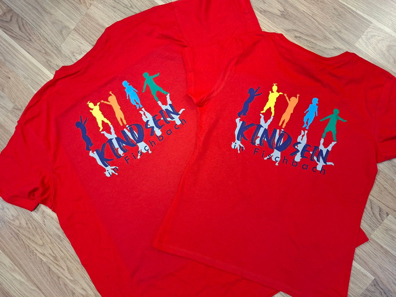 Zwei rote T-Shirts mit dem Wort Kinderse in weißer Schrift liegen auf einer Holzoberfläche. Es gibt verschiedene farbige Silhouetten von Menschen auf den Shirts.