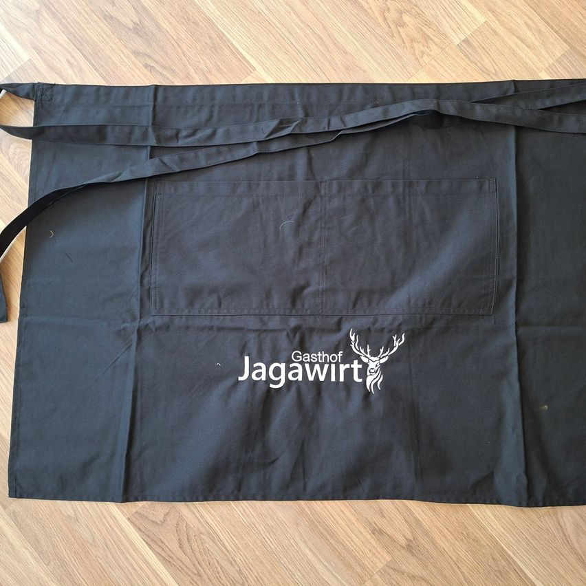 Ein schwarzer Schürze mit dem Text 'Gasthof Jagawirt' und einem Hirschlogo auf einer Holzoberfläche.