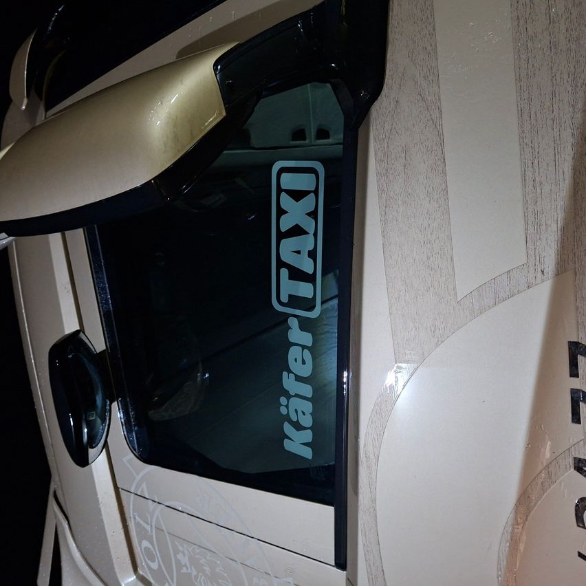 Die Seitenscheibe eines Taxis mit weißem Hintergrund zeigt einen blauen rechteckigen Aufkleber, auf dem in weißen Buchstaben 'Kaffer TAXI' steht. Die Scheibe spiegelt die Umgebung wider, einschließlich des Türgriffs des Taxis und eines Teils des Innenraums.
