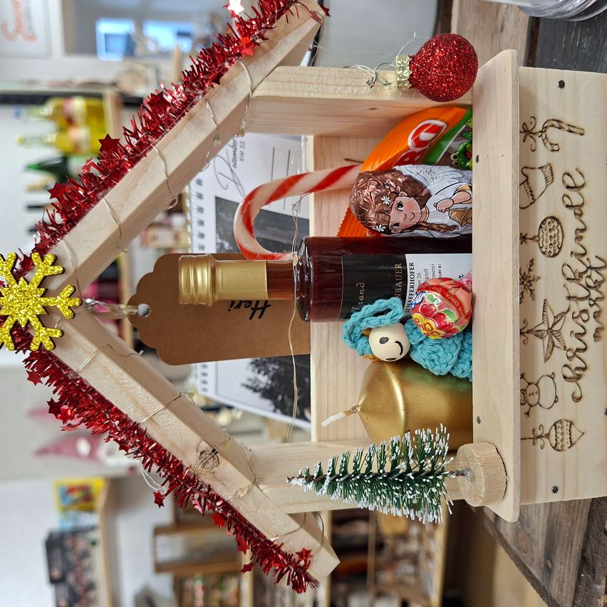 Eine hausförmige Holzbox mit Weihnachtsdekorationen, darunter eine Flasche, eine Zuckerstange, eine Puppe und rotes Tinsel. Die Box hat ein Etikett mit der Aufschrift 'Frohe Weihnachten'. Der Raum ist verschwommen mit einem Regal im Hintergrund.