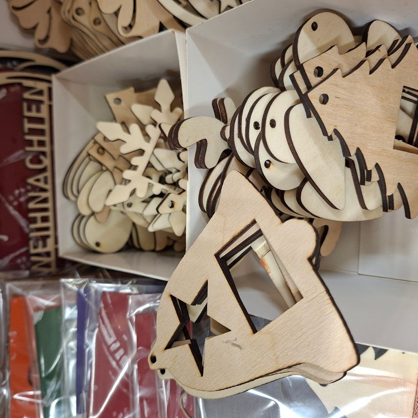 Ein Nahaufnahme-Blick auf verschiedene Holzornamente, darunter Sterne, Schneeflocken und Weihnachtsbäume, die in weißen Boxen und Plastikverpackungen auf Regalen ausgestellt sind.