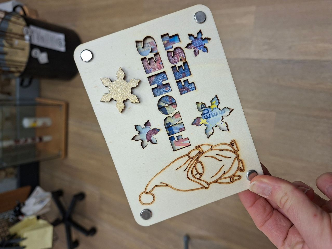 Eine Holzplatte mit dem Wort 'FEST' wird von einer Person gehalten und zeigt Cartoon-Zeichnungen eines Schneemanns und Schneeflocken.