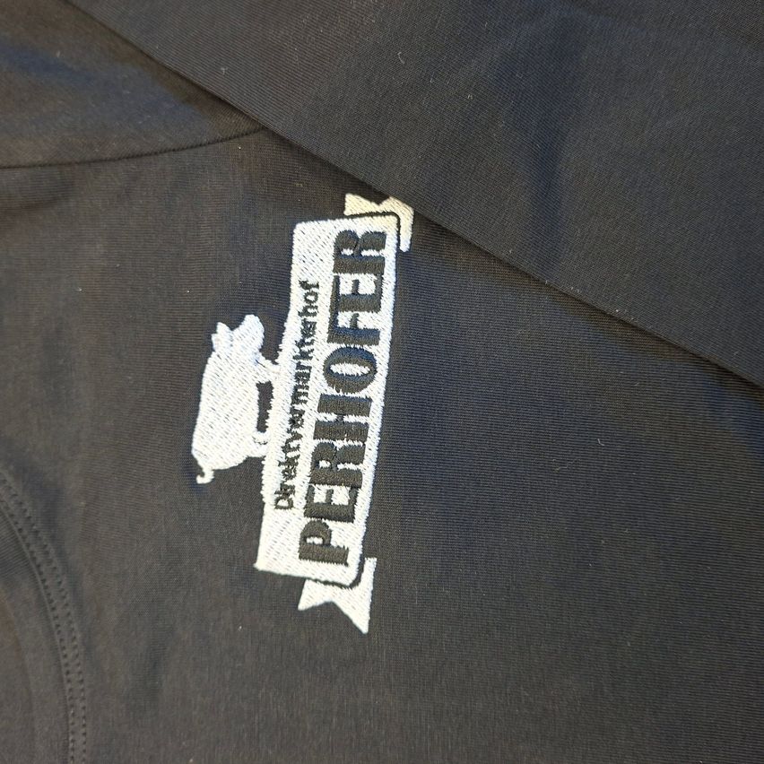 Eine Nahaufnahme eines schwarzen Shirts mit einem weißen gestickten Logo, das 'PERIHOFER' und 'Direktwarenmarkthof' liest.