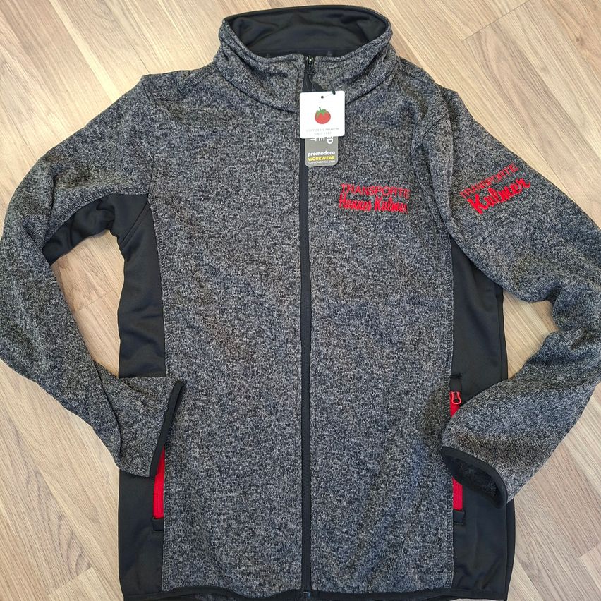 Eine graue und schwarze Jacke mit rotem Text, der 'Transporte Thomas Kalber' lautet, liegt auf einem Holzboden. Sie hat eine Kapuze, einen Reißverschluss und Taschen.