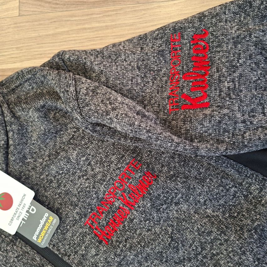 Ein graues Sweatshirt mit roter Stickerei, auf der 'Transporte Kuhlen' und 'Hobby Kuhlen' steht, liegt auf einer Holzoberfläche. Ein Etikett mit einer Tomate und Text ist teilweise sichtbar.