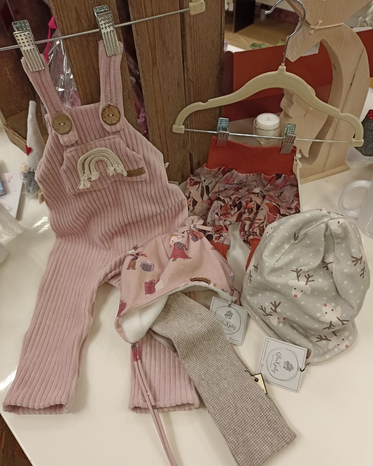 Ein rosa Outfit mit Regenbogen und Knöpfen, ein Blumenrock, eine rosa Mütze und eine graue Mütze mit Hirschen und Schneeflocken.