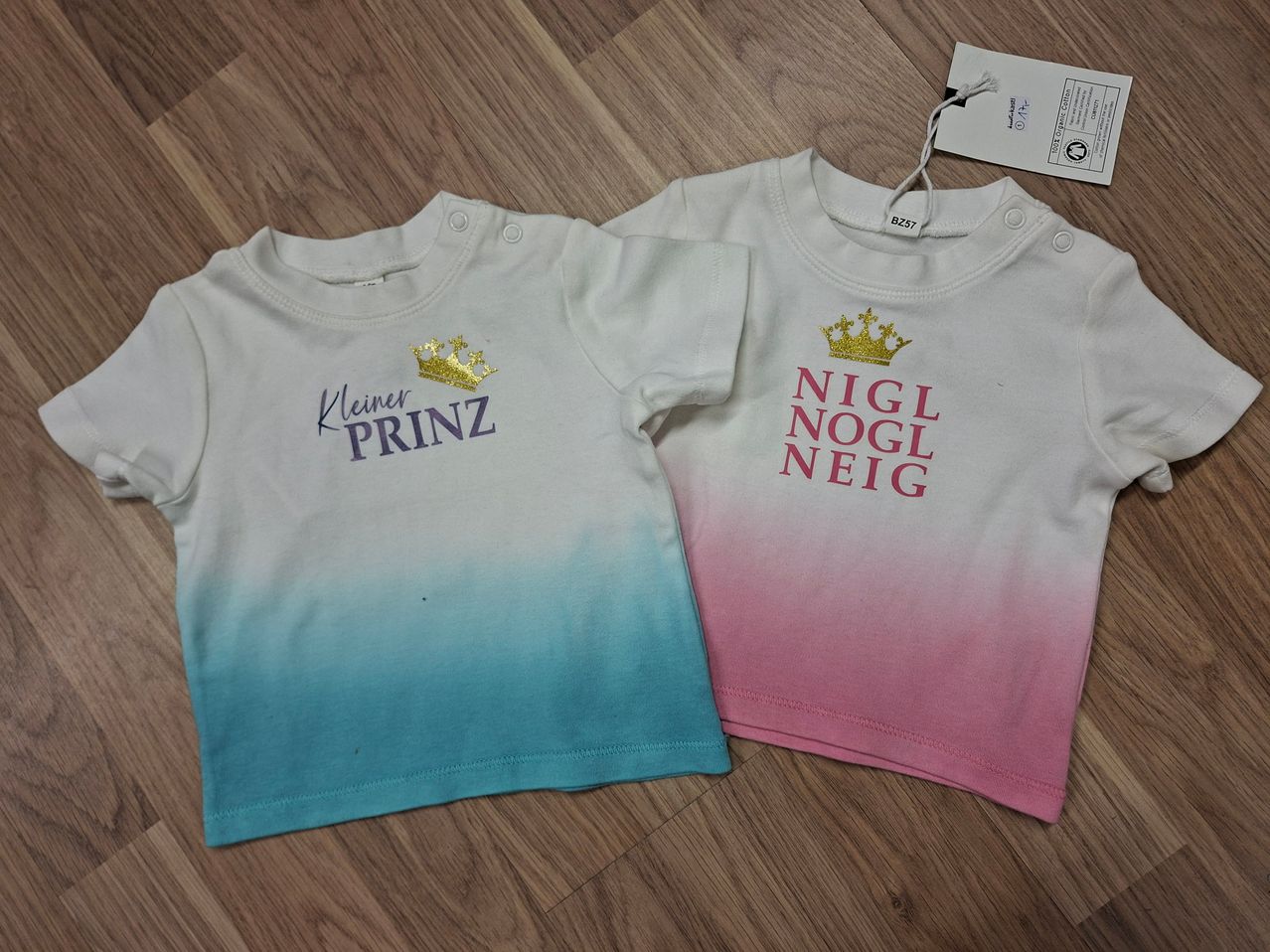 Zwei Baby-T-Shirts auf einem Holzboden. Eines hat einen blau-weißen Farbverlauf mit 'Kleiner Prinz' in Gold. Das andere hat einen rosa-weißen Farbverlauf mit 'Nigl Nogl Neig' in Rot.