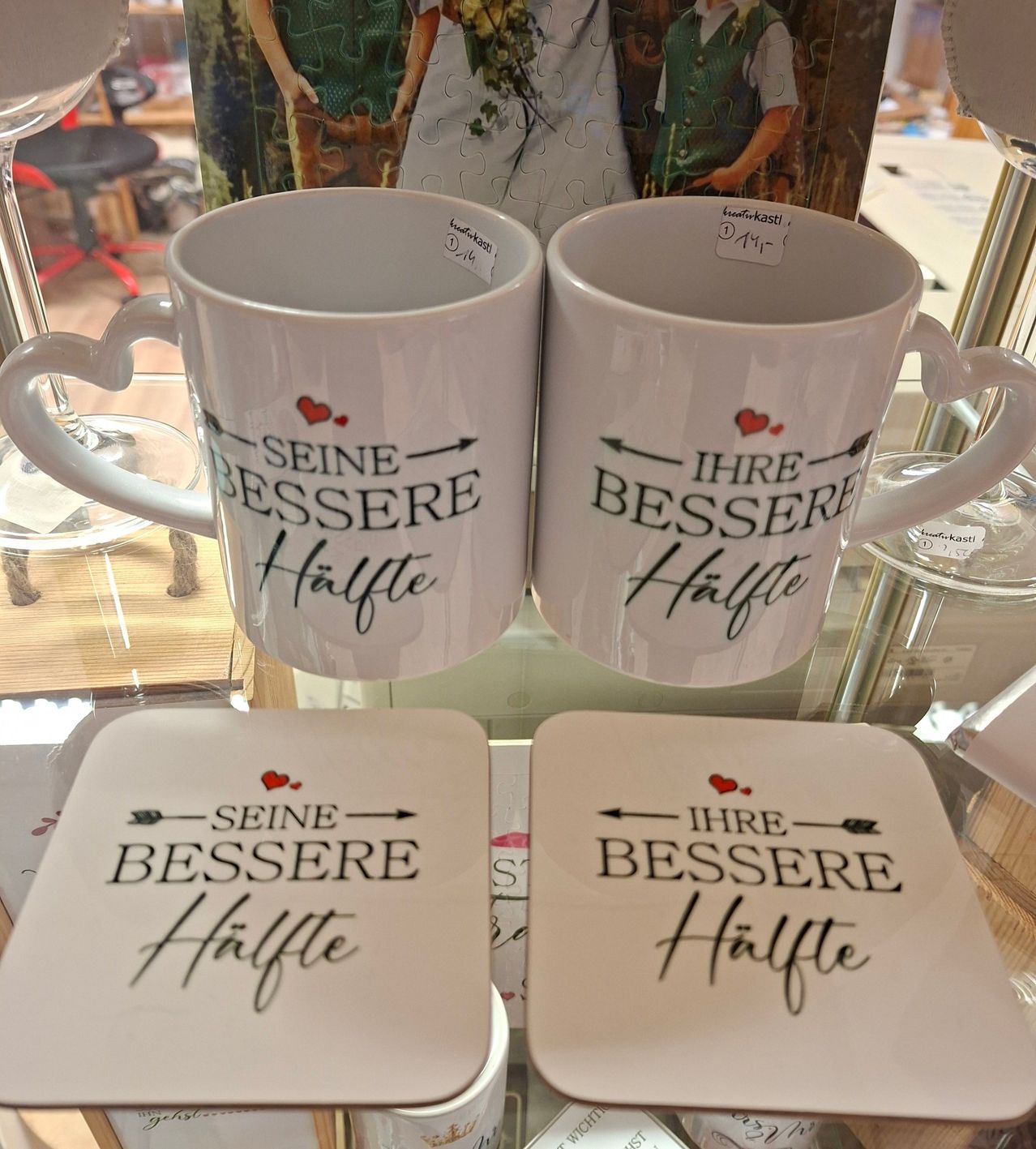 Zwei weiße Tassen und Untersetzer mit den Phrasen 'Seine bessere Hälfte' und 'Ihre bessere Hälfte' auf einem Glasregal ausgestellt.