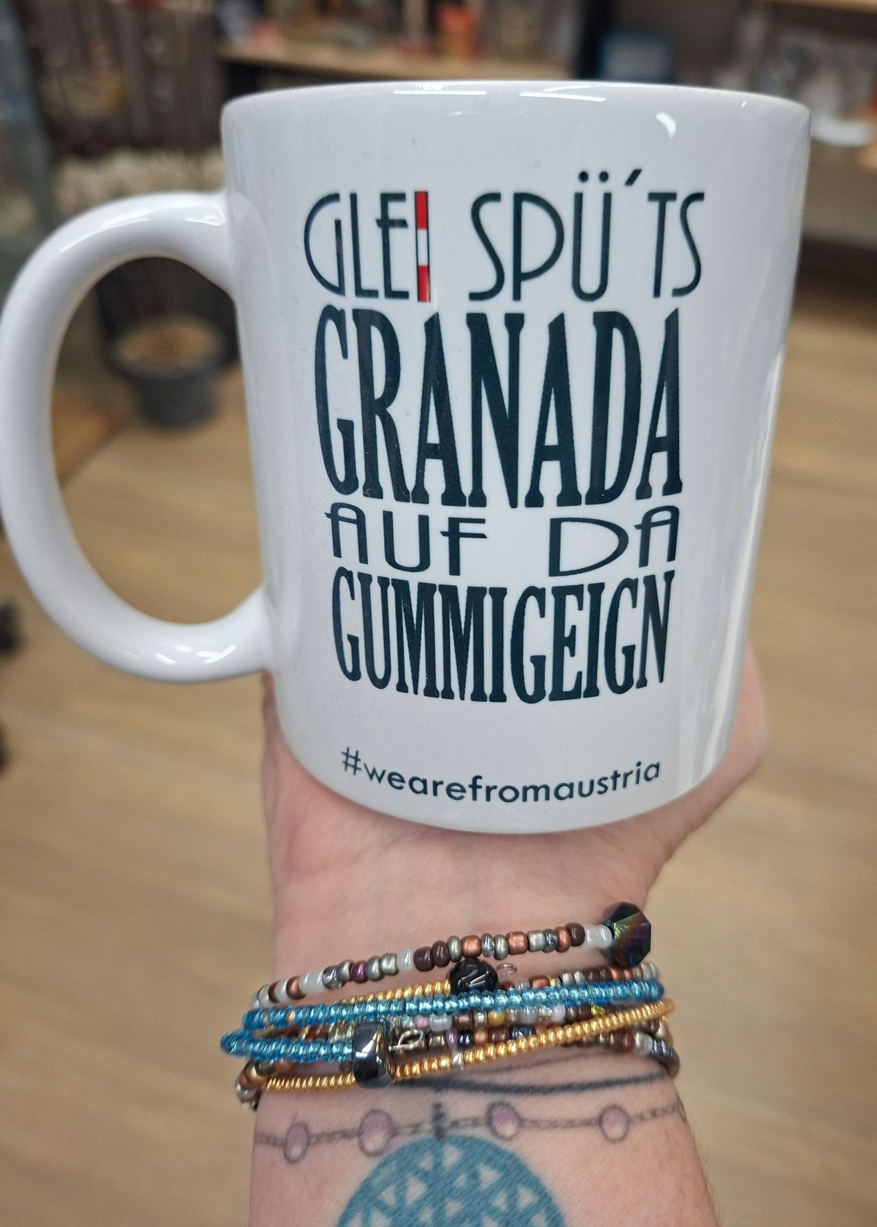 A hand holds a white coffee mug with a colorful design featuring the text 'GLEI SPÜTS GRANADA AUF DA GUMMIGEIGN' and the hashtag #wearefromaustria.