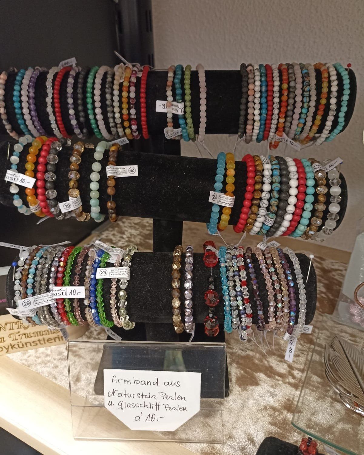 Eine Ausstellung farbiger Perlenarmbänder in verschiedenen Farben. Die Armbänder sind auf einem schwarzen Ständer mit Preisschildern angeordnet. Ein Schild in der Mitte lautet 'Armband aus Kristallsteinen Perlen u. glasschleifif Perlen'.