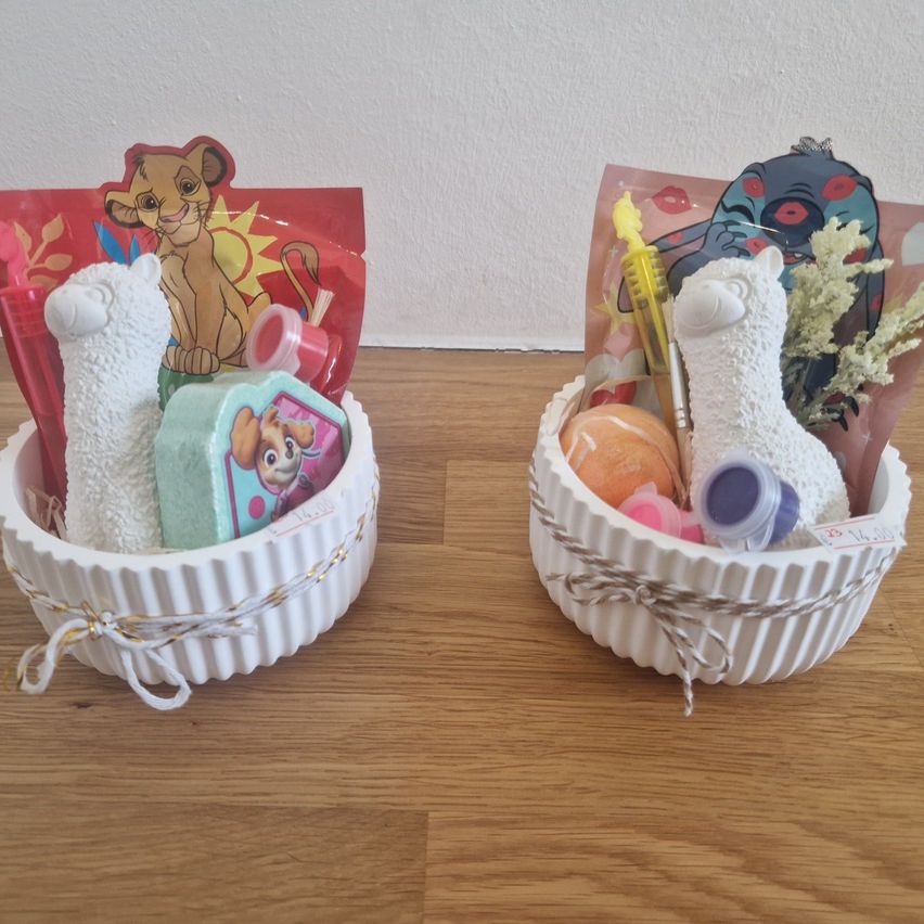 Zwei weiße Keramikkörbe mit Spielzeug und Gegenständen stehen auf einer Holzoberfläche. Jeder Korb hat ein Band. Ein Korb enthält einen Löwenplüsch, eine Zahnbürste und eine Seife. Der andere Korb hat ein Alpaka-Spielzeug, eine Zahnbürste und eine kleine Flasche.