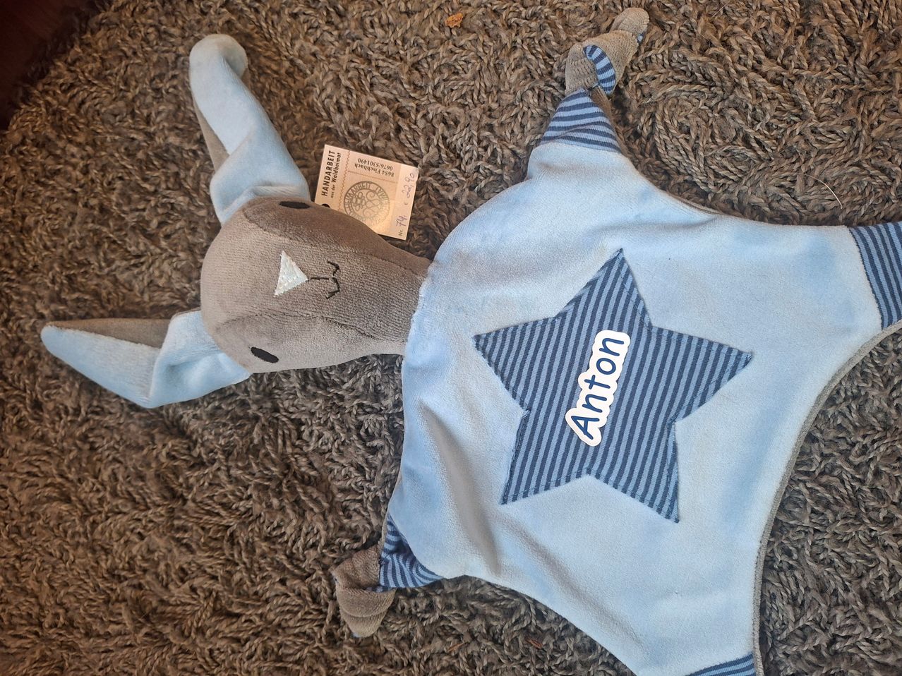 Ein hellblaues Plüsch-Elefantenspielzeug mit einem blauen Stern auf der Brust und dem Namen Anton. Ein altes Ticket liegt neben ihm auf dem Teppich.