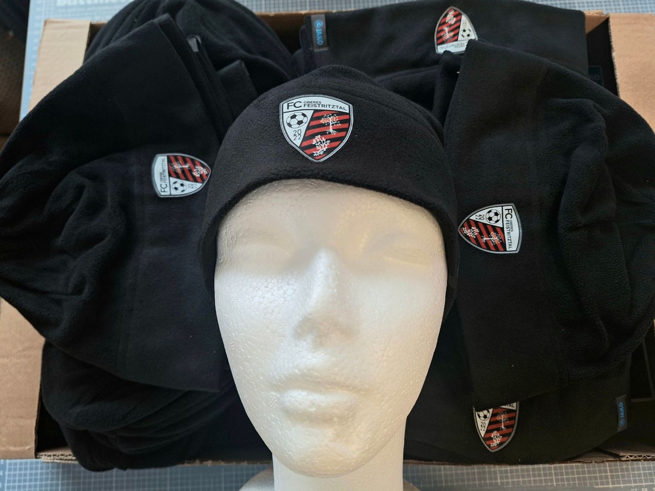 A mannequin head is wearing a black beanie with a red and white logo and number 29. Behind it are other black items, possibly clothes, with the same logo.