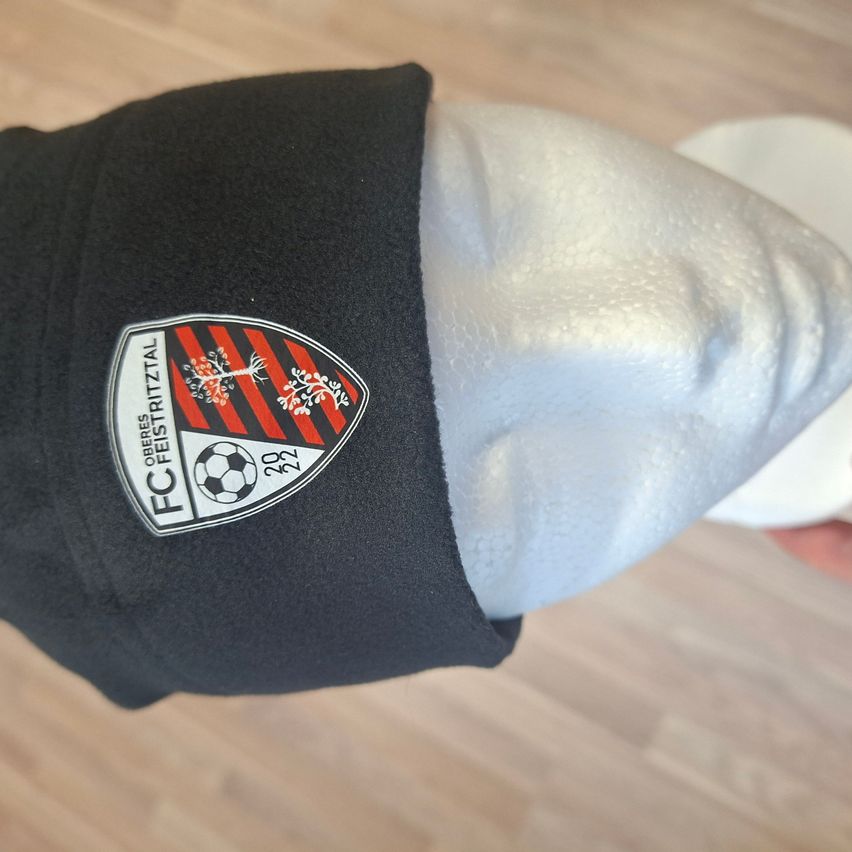 A black hat with a white and red shield logo featuring a soccer ball and a tree, displayed on a mannequin head on a wooden surface.