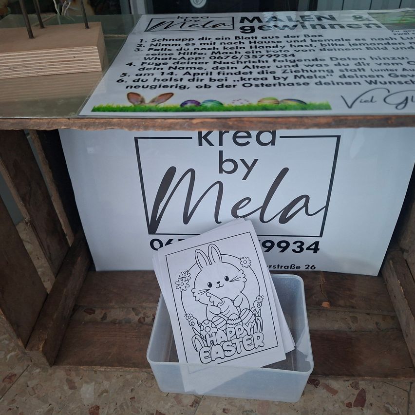 An outdoor wooden table displays a white banner titled 'Krea by Mela'. Below the banner is a clear plastic box containing an Easter coloring book with a bunny illustration.